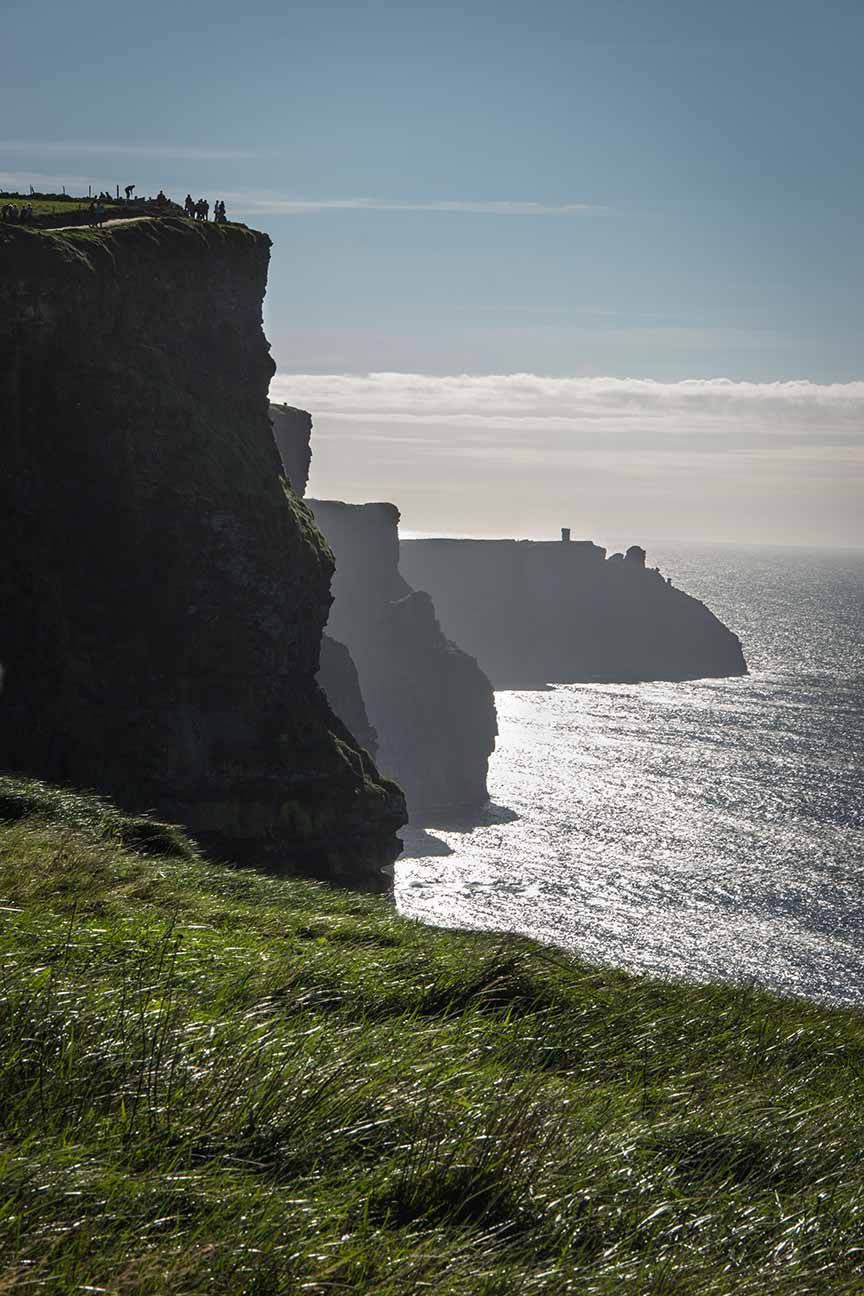 Tourists on the lush green edge of the Cliffs of Moher under a misty sky, overlooking the Atlantic Ocean in Ireland.