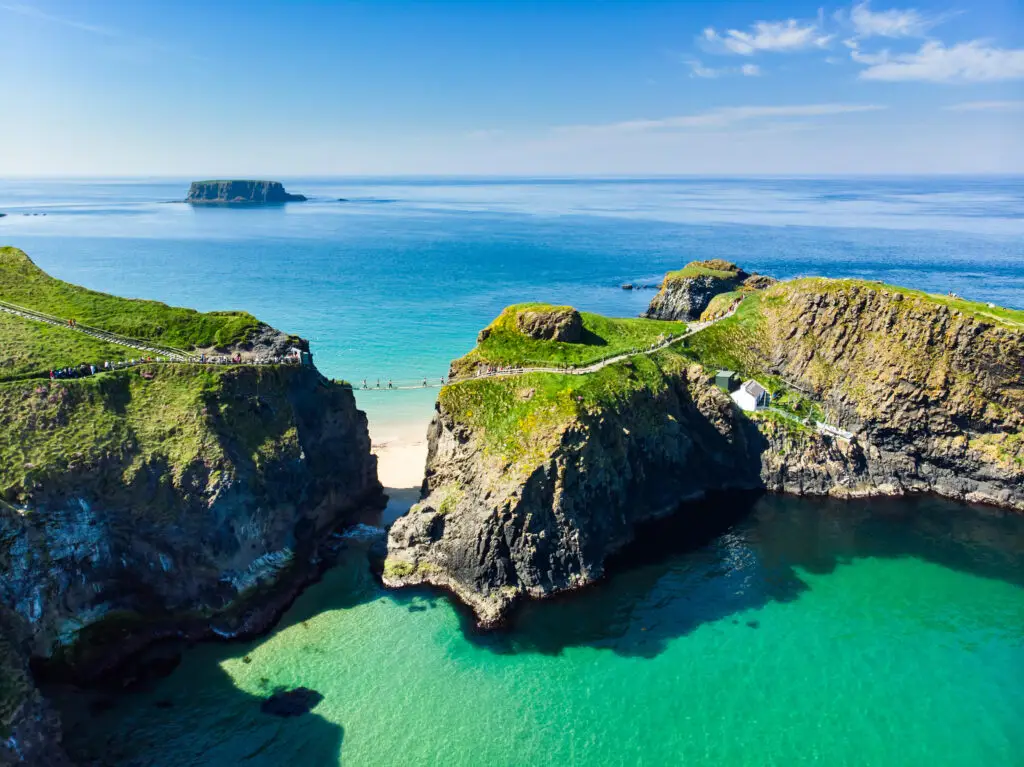 Aerial view of Carrick-a-Rede Rope Bridge in County Antrim, Northern Ireland, connecting the mainland to Carrickarede island, with vibrant turquoise waters and lush cliffs under a clear blue sky.